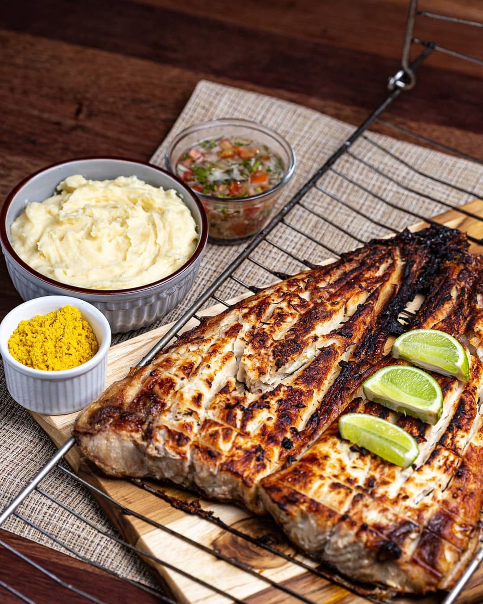 A charred grilled fish fillet with lime wedges, a bowl of salsa and mashed potato