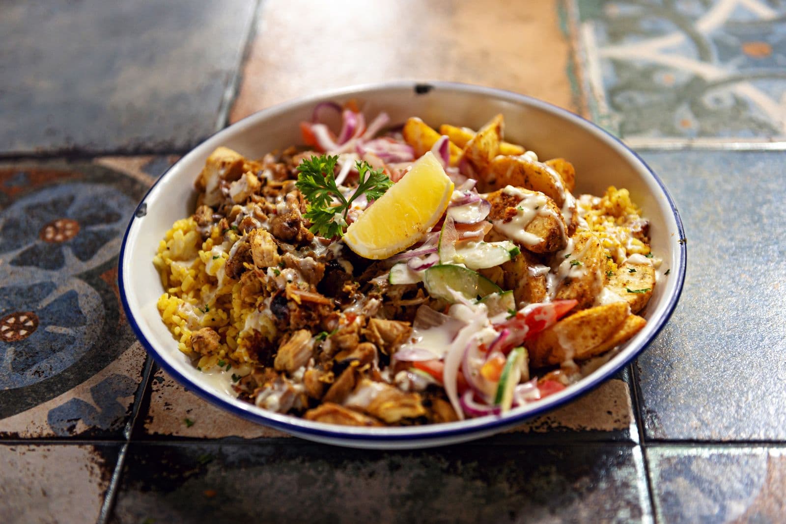 A bowl of spiced chicken over yellow rice with red onion, tomato, cucumber and a lemon wedge